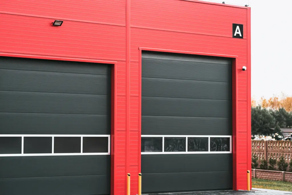 Two dark grey sectional overhead garage doors with window panels on a red metal-clad commercial building marked bay A